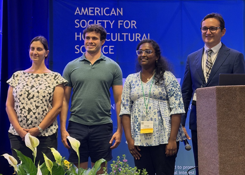 Four individuals stand on a stage in front of a large blue backdrop displaying the words “American Society for Horticultural Science.” Two individuals hold white certificate folders. One person stands at a podium on the right side of the stage. Potted flowering and foliage plants are arranged along the front of the stage. The setting appears to be an awards or recognition ceremony at a professional horticultural science conference.
